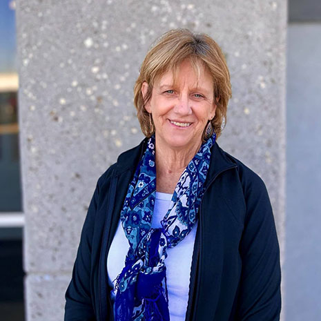 Cindy wearing a white shirt with a dark color cardigan and a blue scarf smiles at the camera. She's standing in front of a gray wall